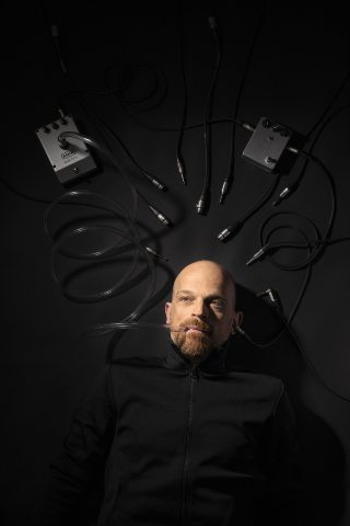 Portrait of Ulrich Troyer in May 2020, with a talkbox and a stompbox, dark atmo, lightspots only on face and effect pedals, photographed by Eva Kelety.